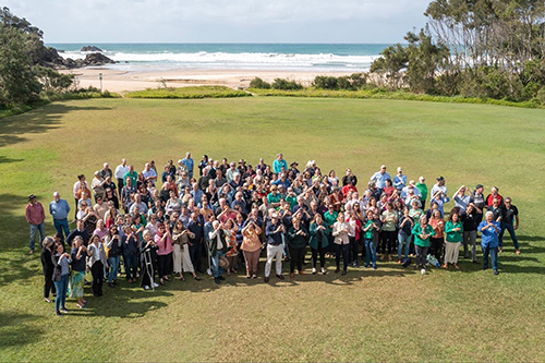A very large group of people standing on a grassy beach front all looking at a birds eye side view smiling and waving up at the camera.