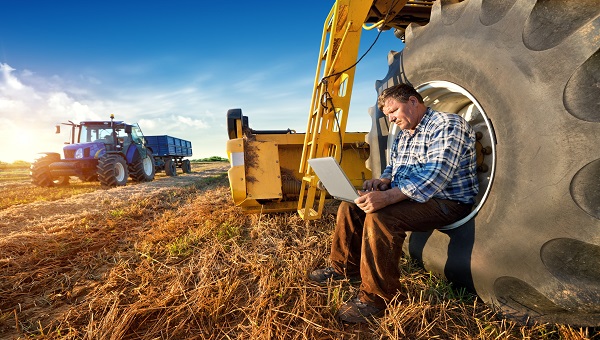 Farmer sits in a tractor wheel with a laptop on his knees, while another tractor drives on a crop in the background under a blue sky. 