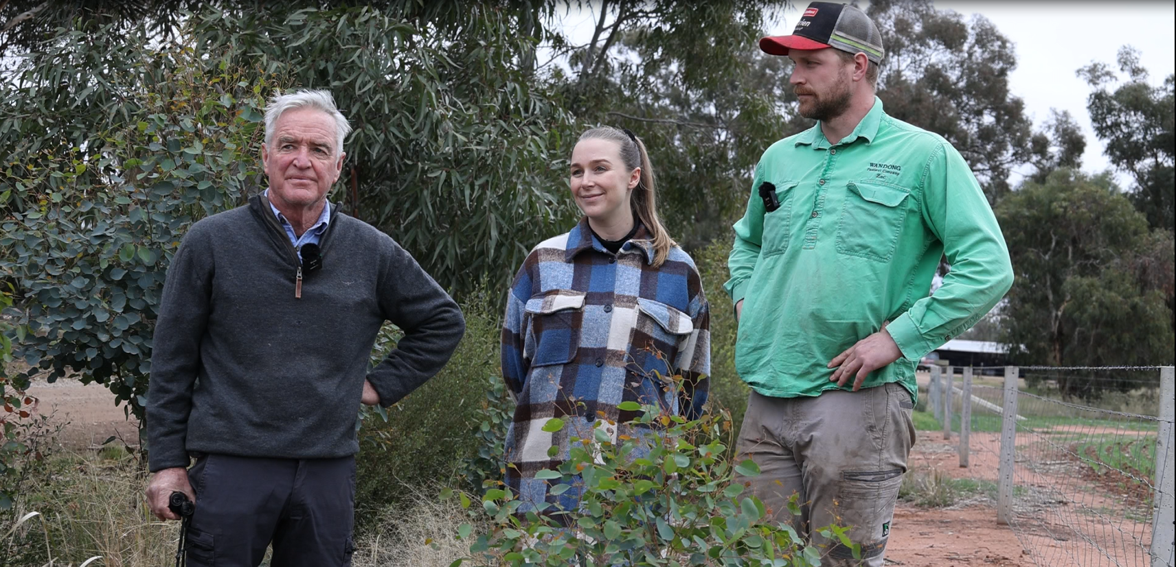 A grey haired man on the left, a lady in a blue checkered shirt in the middle and a man with a green collared shirt on the right, all standing behind a newly planted tree