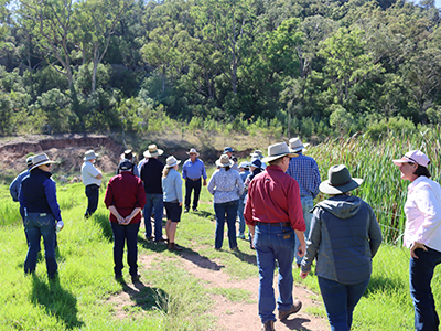 A group of approximately 15 people wearing hats and standing in a paddock next to a river bank.