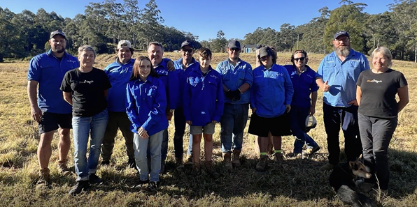 12 people standing in a grassed paddock, facing the camera and smiling. Most appear to be wearing blue work shirts.