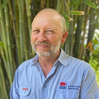 A man in a blue shirt in front of a stand of bamboo. He is wearing a light blue shirt.