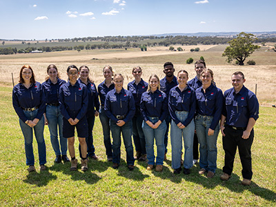 A group of 13 people wearing matching navy shirts, standing in a field and smiling for the camera