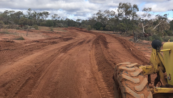 A brown dirt road with a deep ditch running along the right hand side. Heavy machinery sits at the bottom right of teh picture ready to commence work on fixing the road erosion 