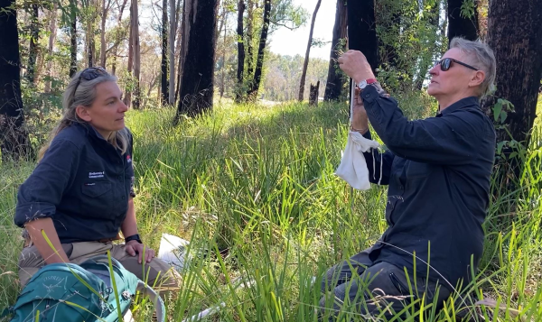 Two people in uniform sitting facing each other in long grass. One person is holding a measuring device with a small cloth bag, reading off a metre while the other watches. 