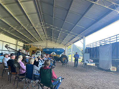 A group of people sitting on camp chairs under a large metal shed structure. A person is standing in front of the crowd next to a tresle table and in the background is a truck and yellow tractor. 